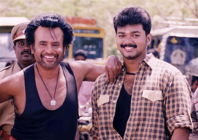 A vintage behind-the-scenes photograph of Tamil cinema actors Rajinikanth and Vijay smiling together on a film set, with Rajinikanth putting his arm around Vijay's shoulder